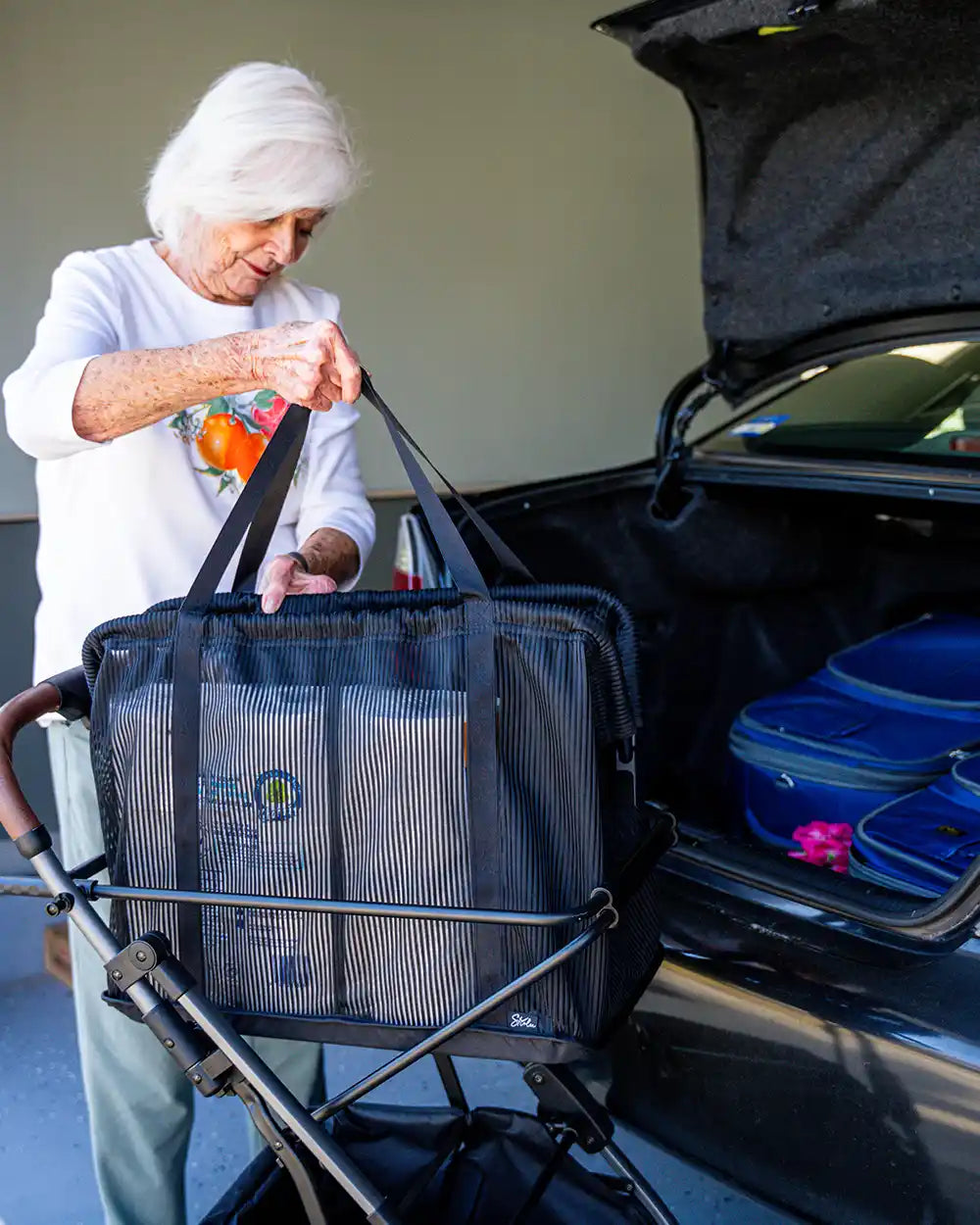 Frau hält die Tragegriffe eines gefüllten Personal Shopping Bag auf einem Strolee-Wagen neben einem offenen Kofferraum.